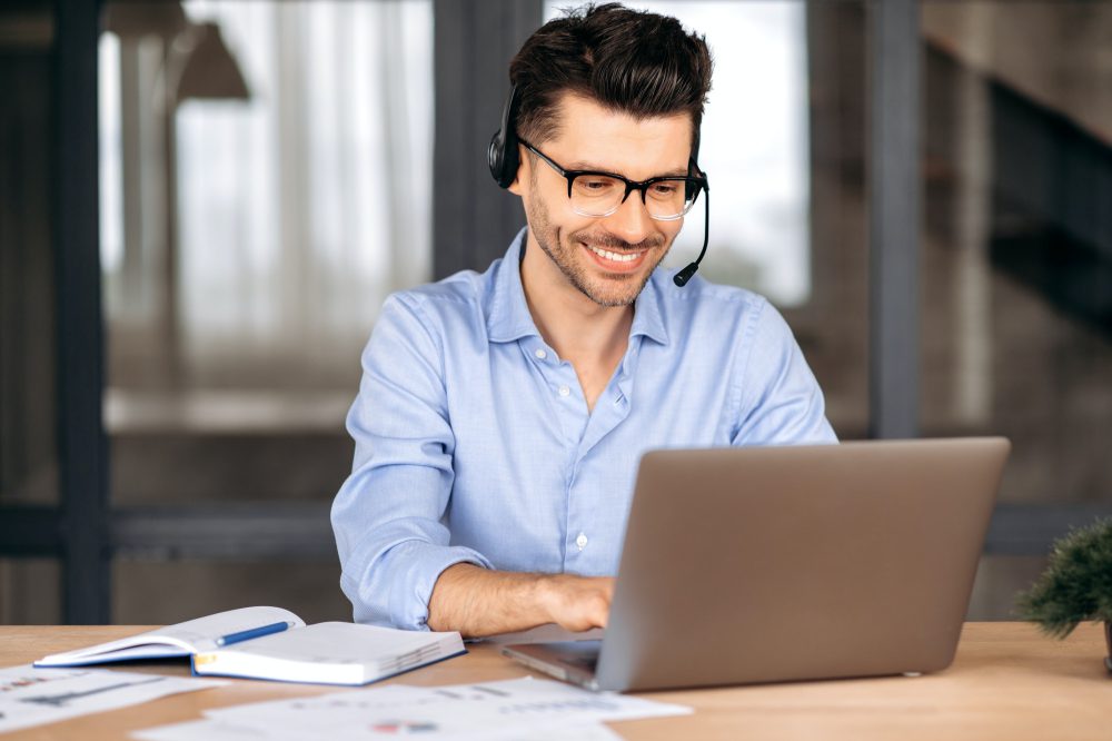 Friendly man using laptop, chatting with client online, typing on keyboard, smiling