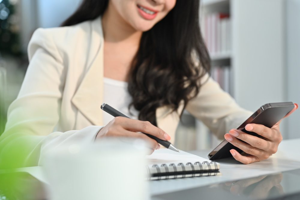Smiling young businesswoman holding phone making important notes, planning daily.